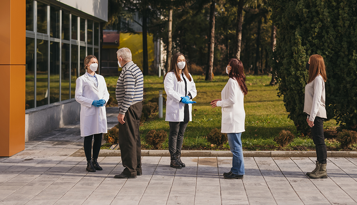 Group of senior, mid and young adult people with protective face masks standing in a line in front of a hospital and waiting for coronavirus vaccination, while young female doctors giving them explanations and directions