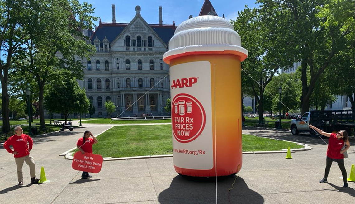 a a r p volunteers hold up a giant inflatable pill bottle with signs advocating for fair drug prices now