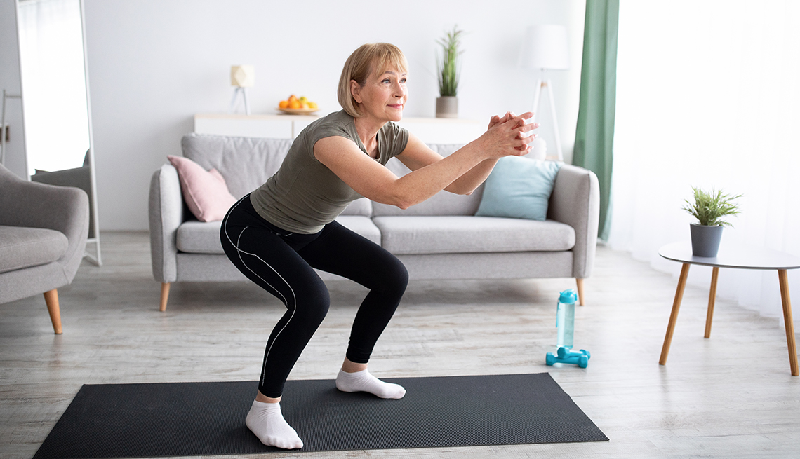 Positive senior lady doing squats on domestic workout in living room, free space. Fit mature woman leading active lifestyle, losing weight, staying in good shape at home during coronavirus quarantine